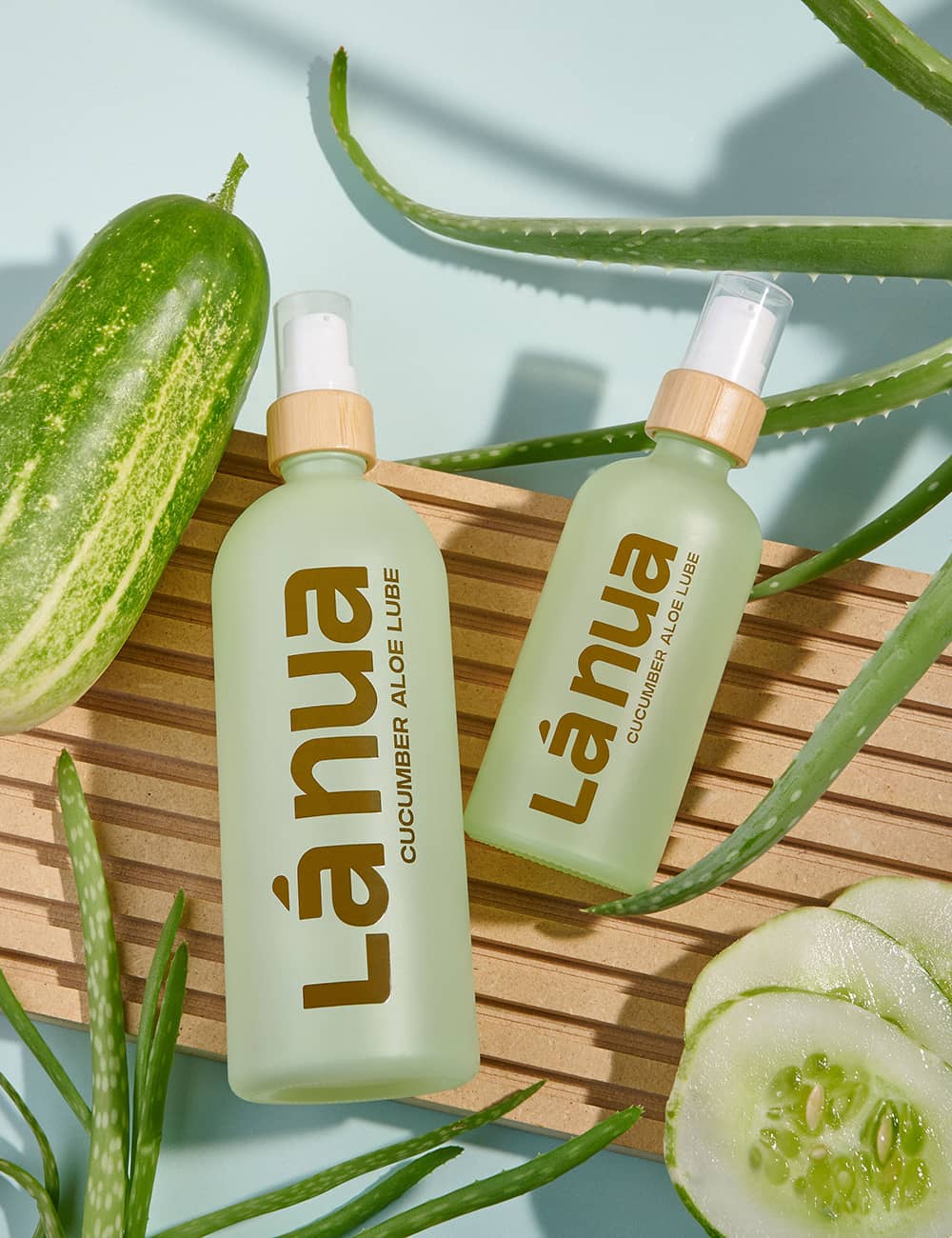Two bottles of La nua cucumber aloe flavored lube with cucumbers and aloe vera leaves on a wooden surface.