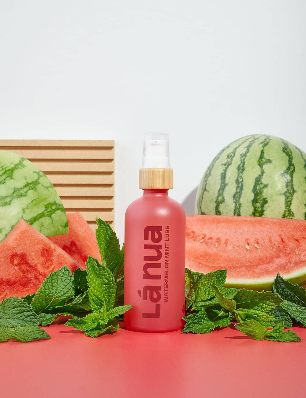 Pink flavored lube bottle with a gold cap labeled 'la nua' surrounded by watermelon slices and leaves on a red surface.